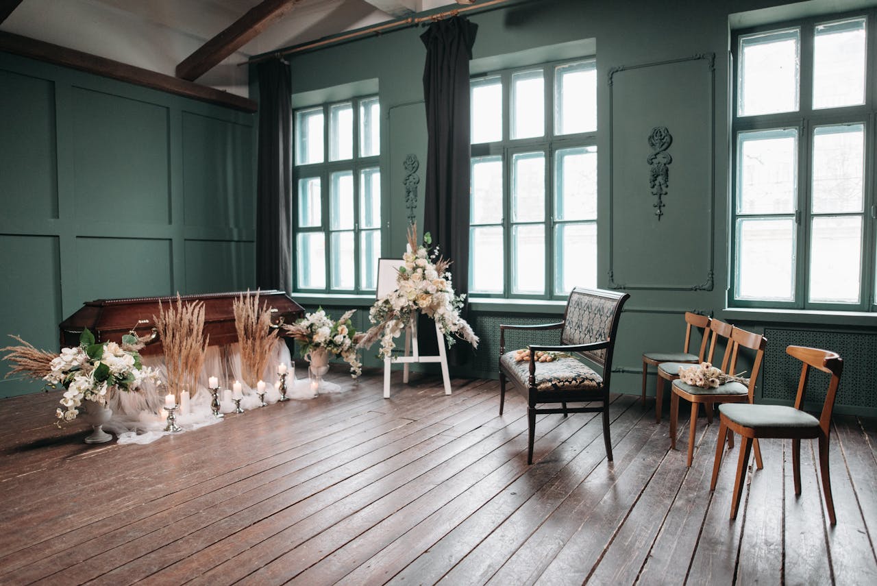 A serene funeral setup with a wooden coffin, chairs, and floral arrangements in a green room.