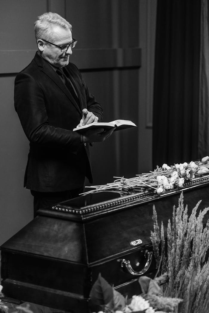 Black and white image of a pastor reading the Bible at a funeral service with a coffin and flowers.